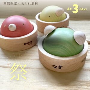 名入れ無料キャンペーン中の木のガラガラ。3色の動物型おもちゃ（赤・黄・緑）が並び、手前の緑のかえるに“なぎ”と名入れされた様子