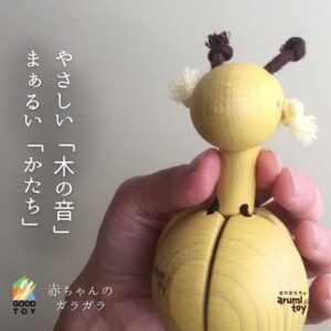 手に持たれた黄色い木製のキリン型ガラガラ。やさしい木の音とまあるいかたちが特徴で、グッド・トイ認定ロゴと“赤ちゃんのガラガラ”の文字入り