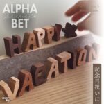 「HAPPY VACATION」と並べられた木製アルファベットブロック。ウォルナットやメイプルなど異なる木材で作られた文字が、温もりあるナチュラルな質感で表現されている。手が文字を動かす様子と、「記念日祝いに」の文字入り。
