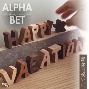 「HAPPY VACATION」と並べられた木製アルファベットブロック。ウォルナットやメイプルなど異なる木材で作られた文字が、温もりあるナチュラルな質感で表現されている。手が文字を動かす様子と、「記念日祝いに」の文字入り。