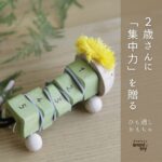 天然木で作られた毛糸のたてがみを持つイモムシ型のひも通しおもちゃ。ブロック部分に1から5の数字が刻まれ、ひもが通されている。背景は木のテーブルとグリーンの植物。右側には「2歳さんに『集中力』を贈る ひも通しおもちゃ」と縦書きの文字と「arumitoy」のロゴが配置されている。