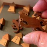 木でつくられたやもりのマグネットを指先で持ち上げている様子。背景には家の形をした小さな木製オブジェが並び、工房のあたたかな雰囲気が伝わる写真。